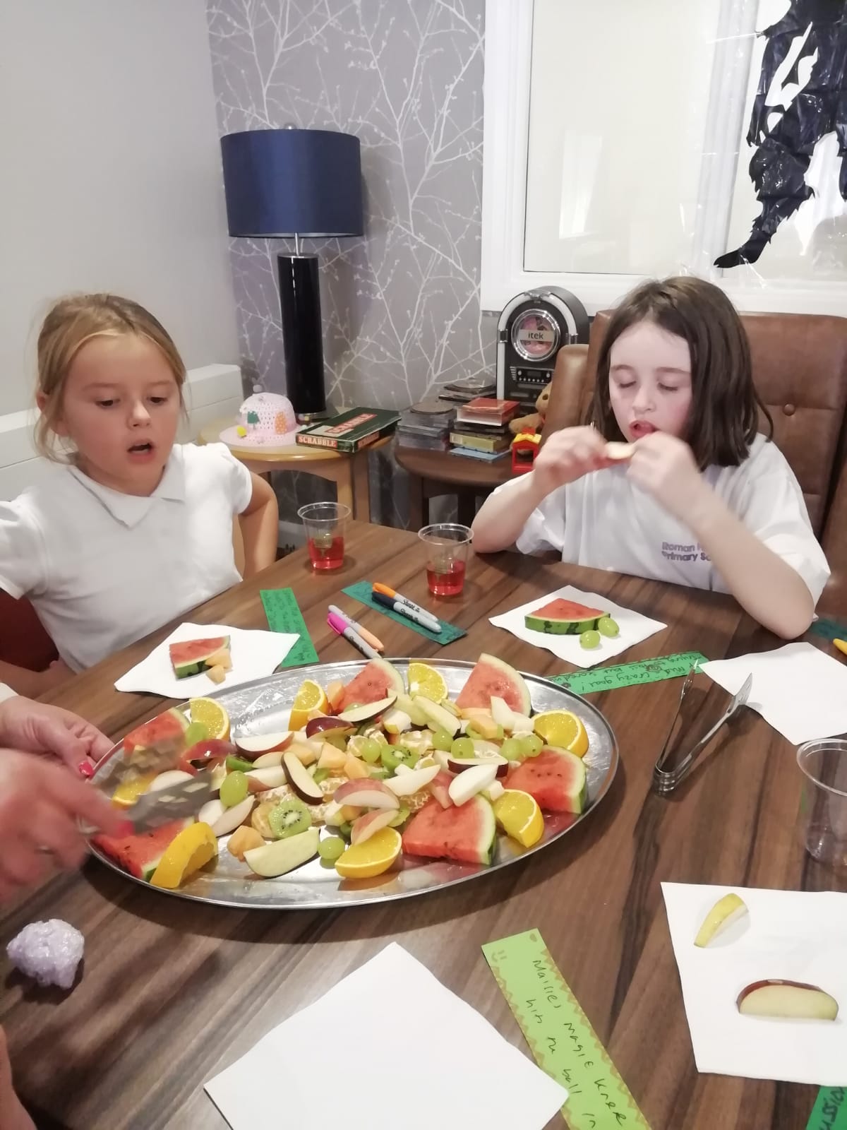 Two school girls, wearing white T-shirts, seated at a table with a large silver platter of fruit in front of them