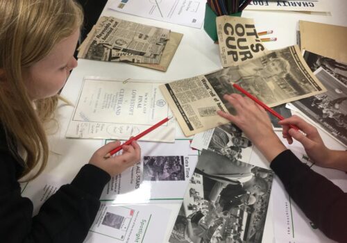 A table top covered in old press cuttings with overhead view of two school pupils, each holding a red pencil, pointing to an image