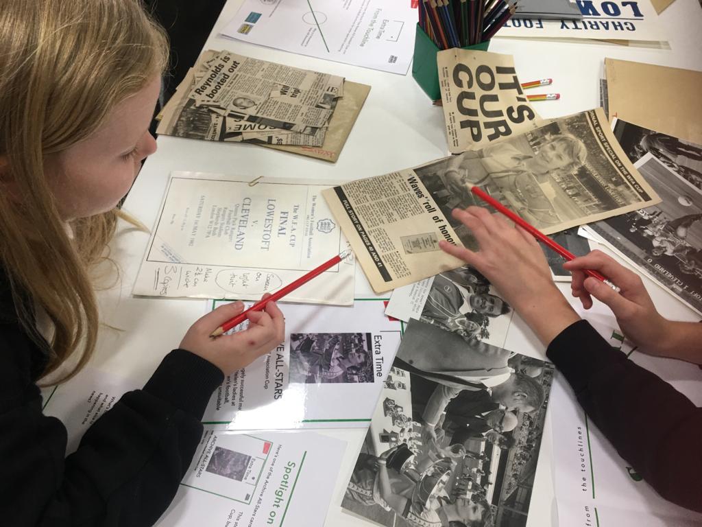 A table top covered in old press cuttings with overhead view of two school pupils, each holding a red pencil, pointing to an image