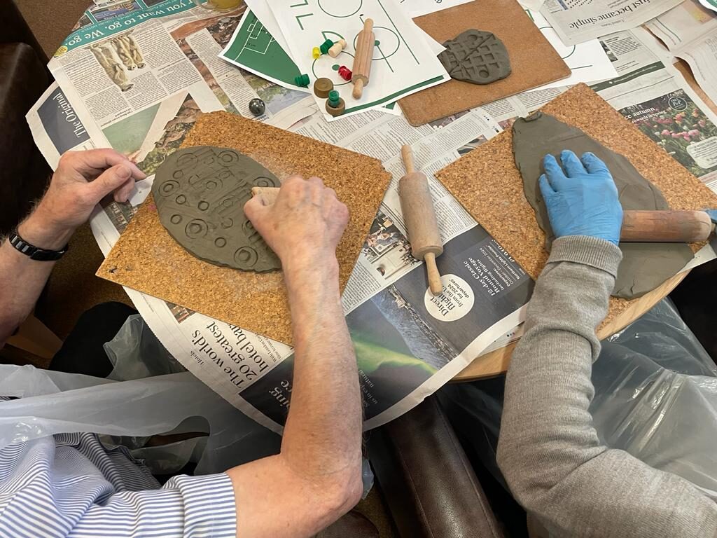 An overhead view of two pairs of adult hands, one pair in lightweight blue gloves, working on slabs of clay in front of them