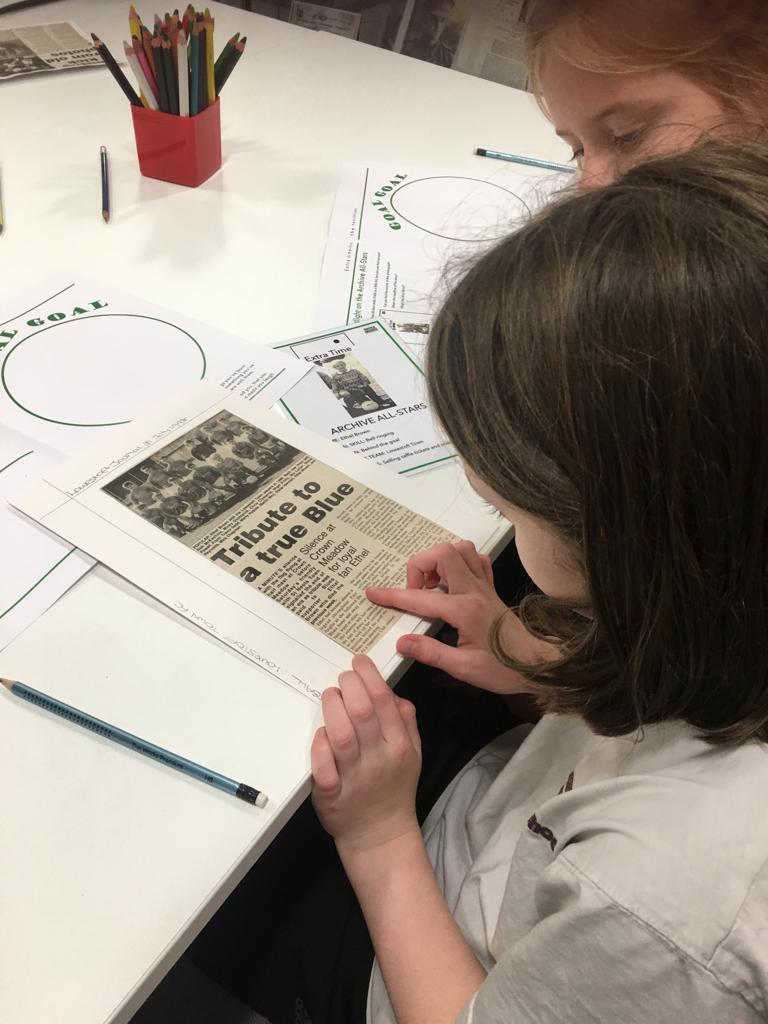 Close up of two school children seated at a table on which is a pot of coloured pencils and some papers. They are reading a press cutting and pointing to a particular line