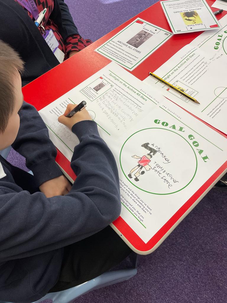 A young boy writes something next to a hand drawn image of a footballer in a green circle on white paper.