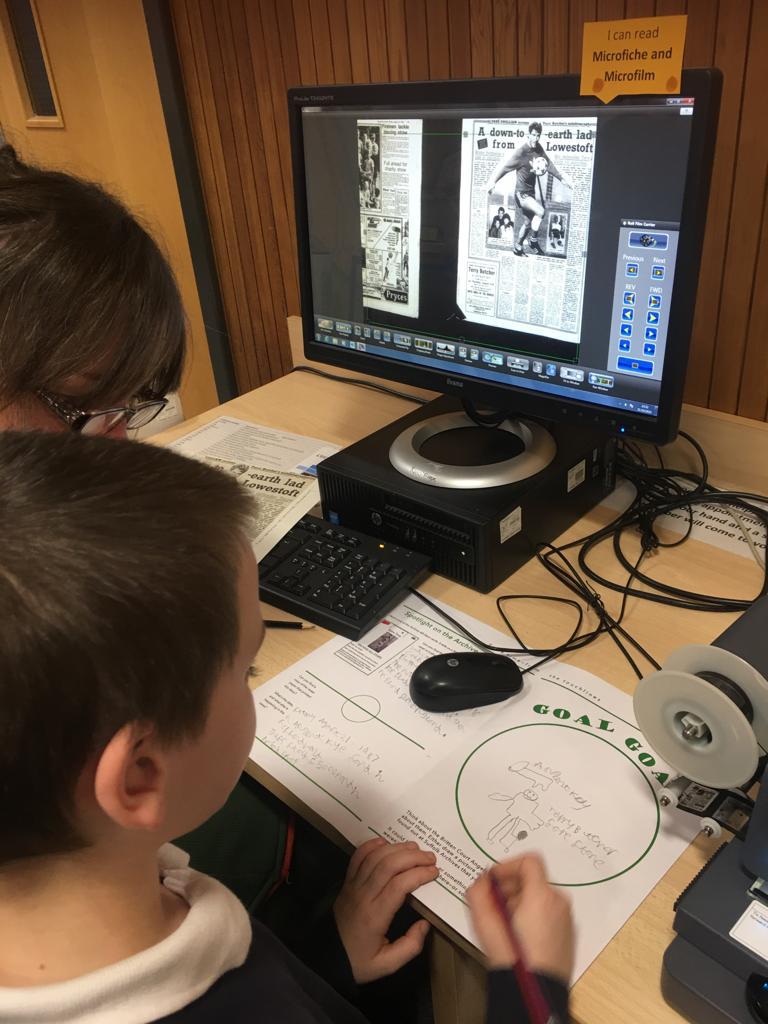 A young boy sits in front of a screen, using microfiche to look at old press cuttings