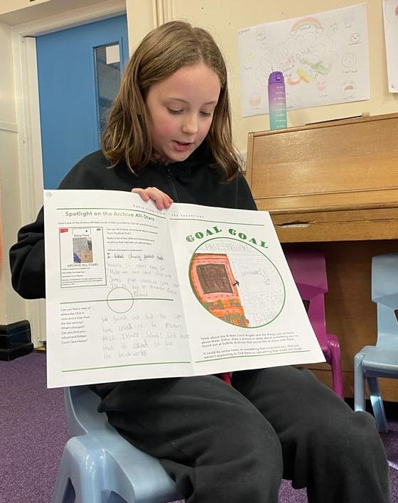 A child dressed in dark school uniform is seated and is obviously showing a worksheet to others (unseen) in the room