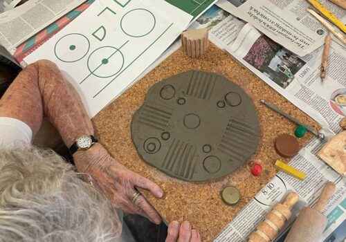 A grey disc of soft clay patterned with small circles and lines, with an array of mark making items beside it