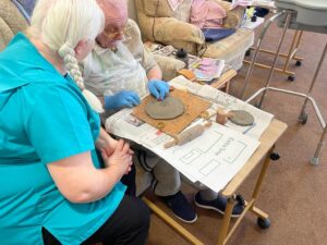 A gentleman is seated with a small table in front of him on which he works on a soft clay disc, watched by a member of the care staff