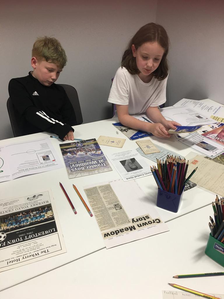 A school boy and girl sit side by side in front of a table with press cuttings and pots of coloured pencils