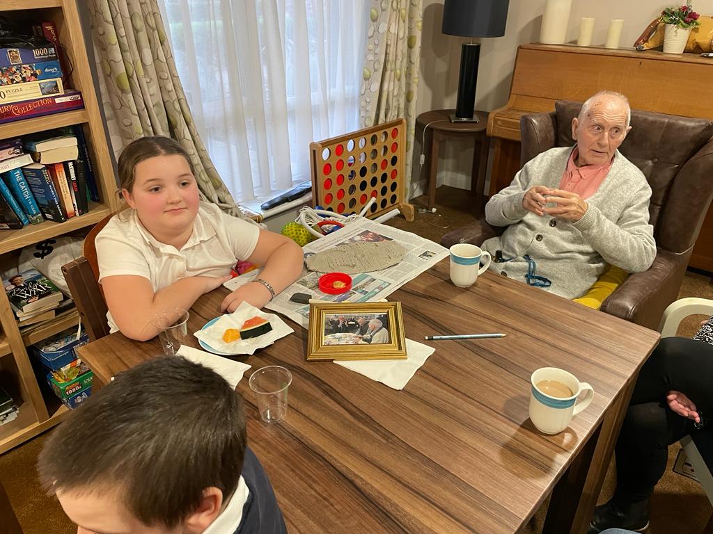 A gentleman is seated at a table, beside a school girl. On the table are mugs of tea, some fruit and a framed photograph that clearly shows the seated gentleman