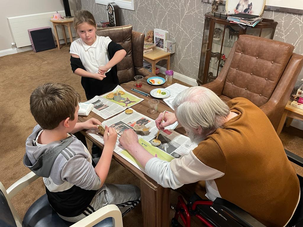A table top covered in newspaper with two children and an adult seated around it, painting small objects. The girl is twisting the top off a paint pot