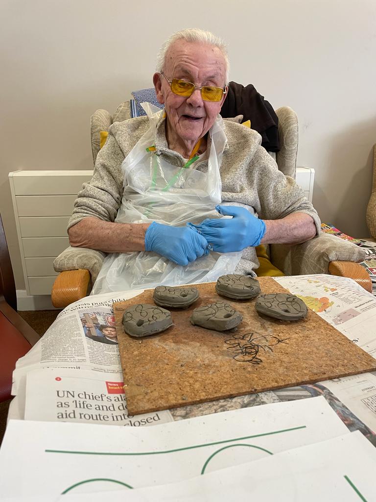 A gentleman seated, with a white plastic apron, in front of him a cork board on a table, with five small discs cut from his original piece of worked clay