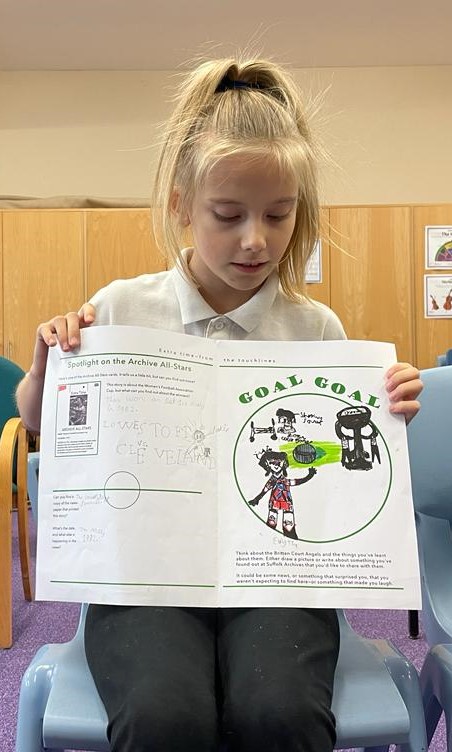 A school girl, with hair bunched up on top of her head, shows her worksheet with pictures of footballers