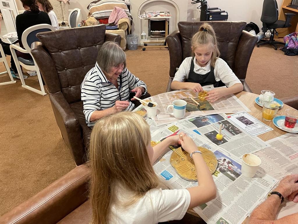 An adult and two children seated at a table covered in newspaper, mugs of tea and plates, painting clay discs gold