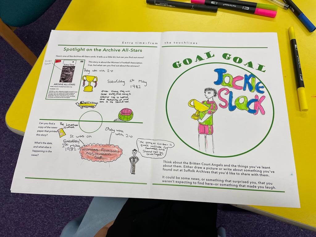 A worksheet on a yellow table with coloured pens around it used to create pictures of a footballer holding a large gold cup