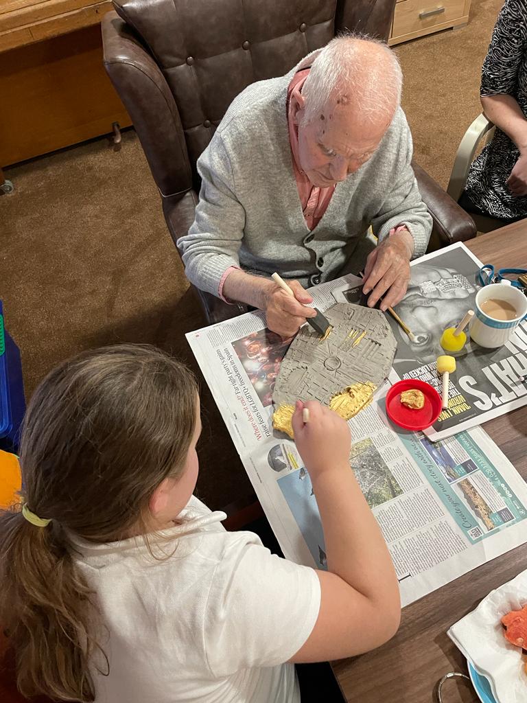 An overhead view of a gentleman and a school girl working on the same clay plaque, painting it gold