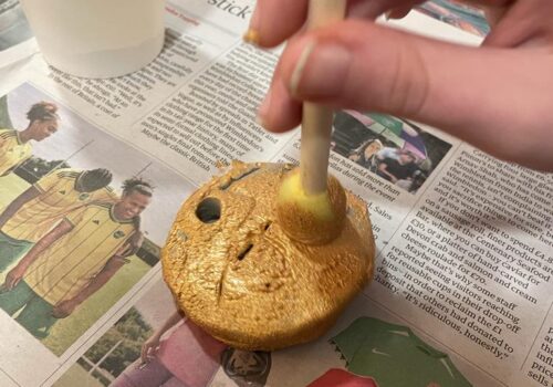 Close up of a hand holding a small stick with sponge, applying gold paint to a small disc with a hole pierced in it.
