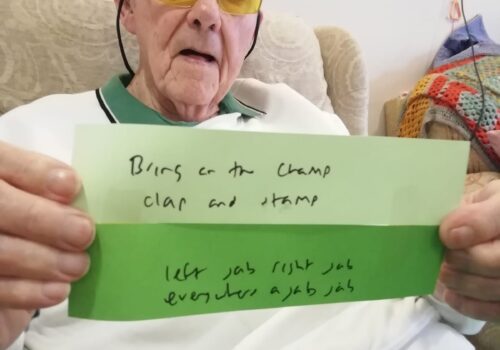 Gentleman seated in armchair holding up to strips of paper with handwritten notes saying Bring on the Champ, Clap and Stamp, Left jab, right jab, everywhere a jab jab