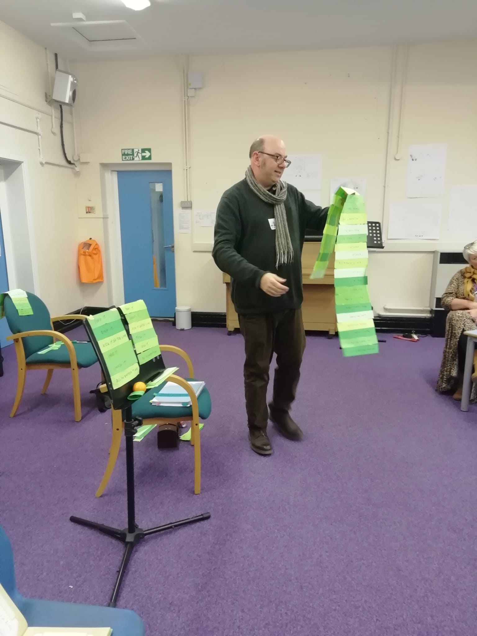 Gentlman with dark jumper and long scarf round his neck stands in class room reading off a long strip of green paper