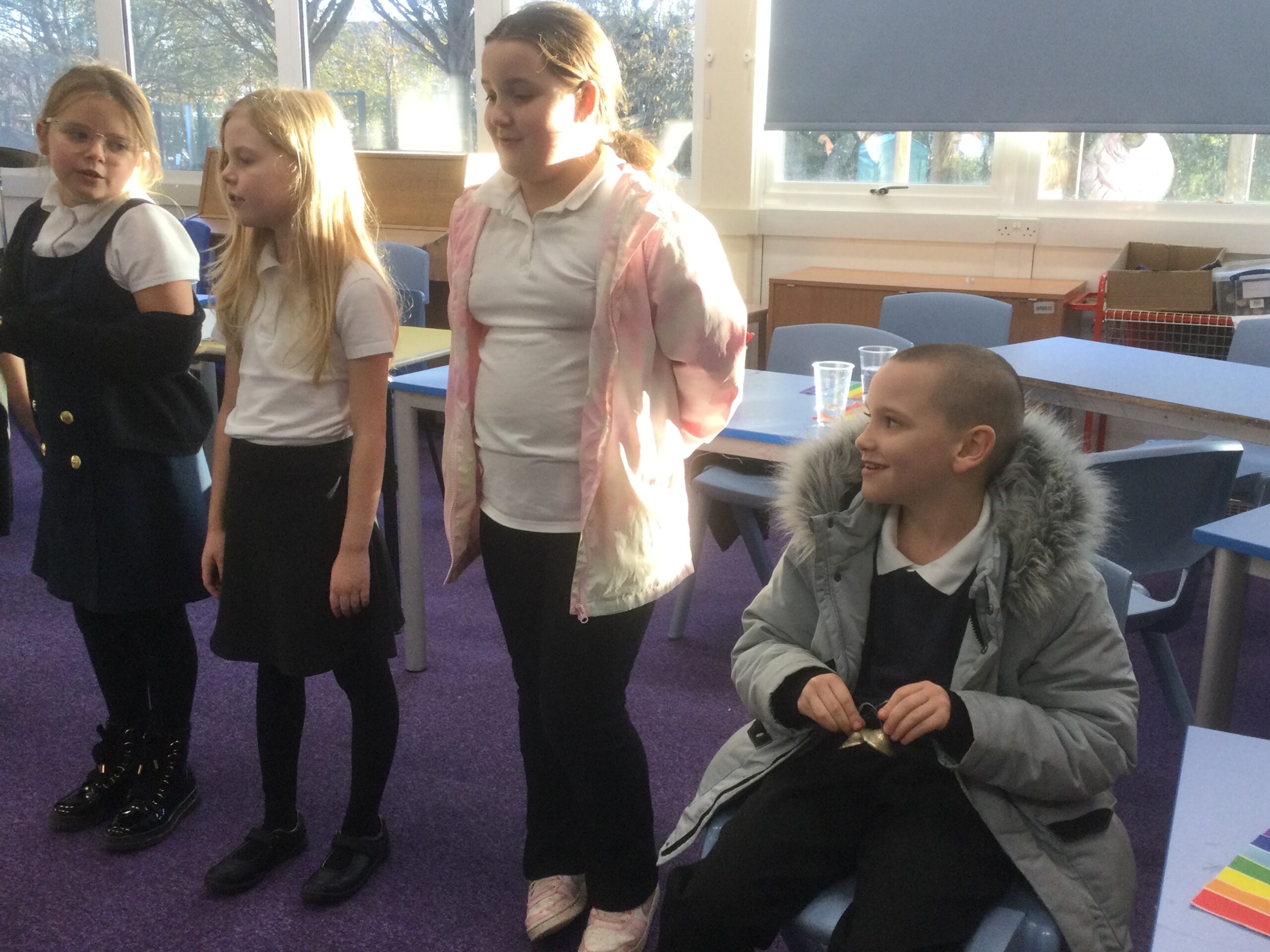 A row of three school girls standing, one wearing a pink cardigan, with a young boy, wearing a grey coat with a fur trimmed hood, seated, holding a pair of meditation bells