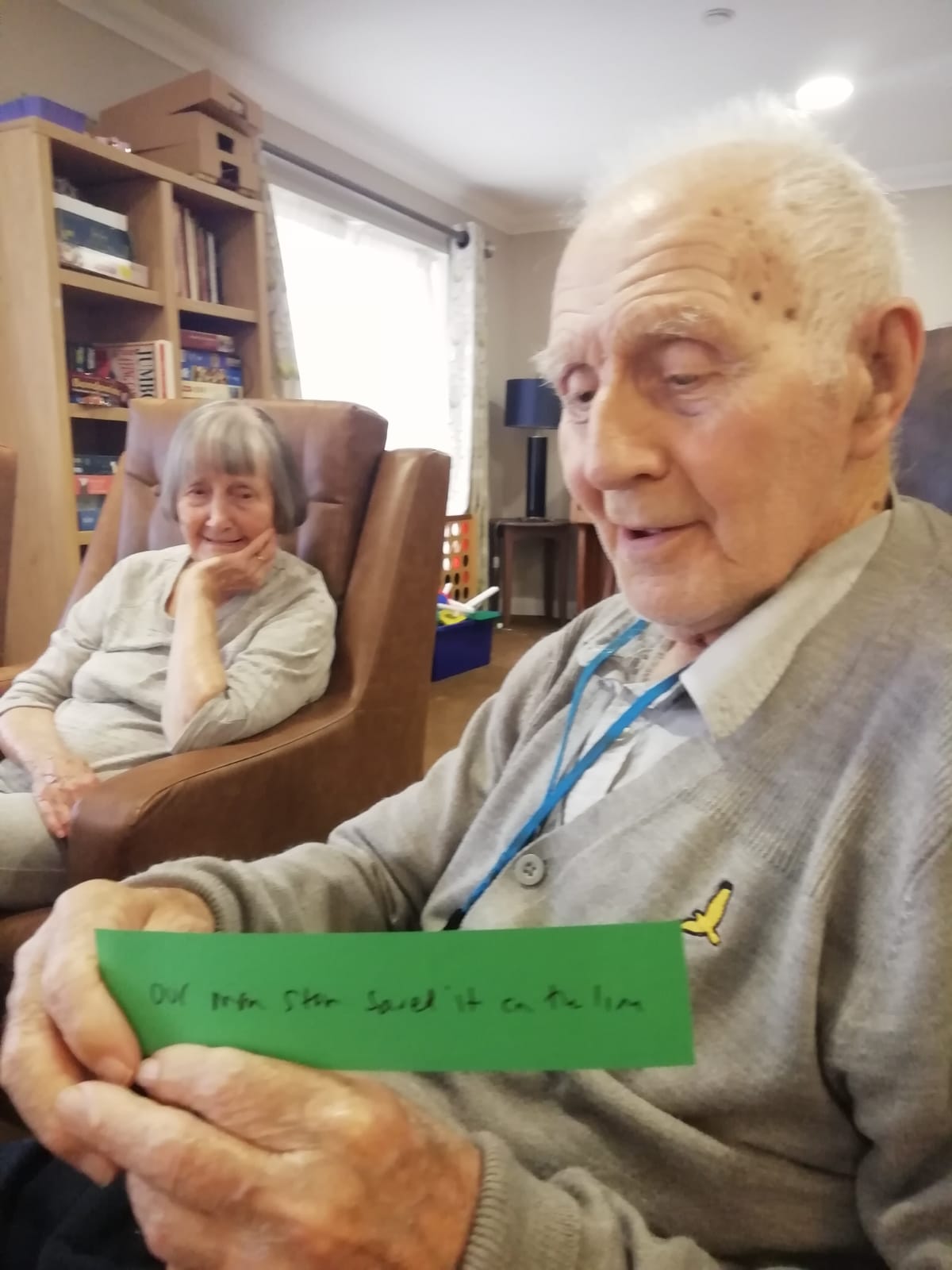 A gentleman seated, holding a strip of green paper, with woman seated in brown armchair looking on