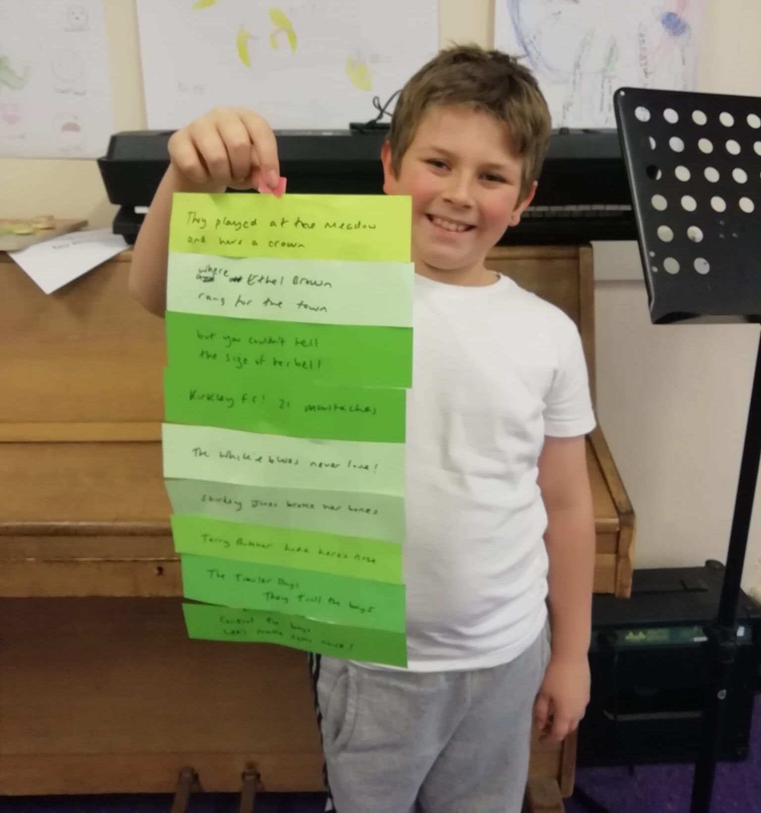 A young boy wearing white T-shirt and grey jogging trousers, holding up a poem written on strips of green paper