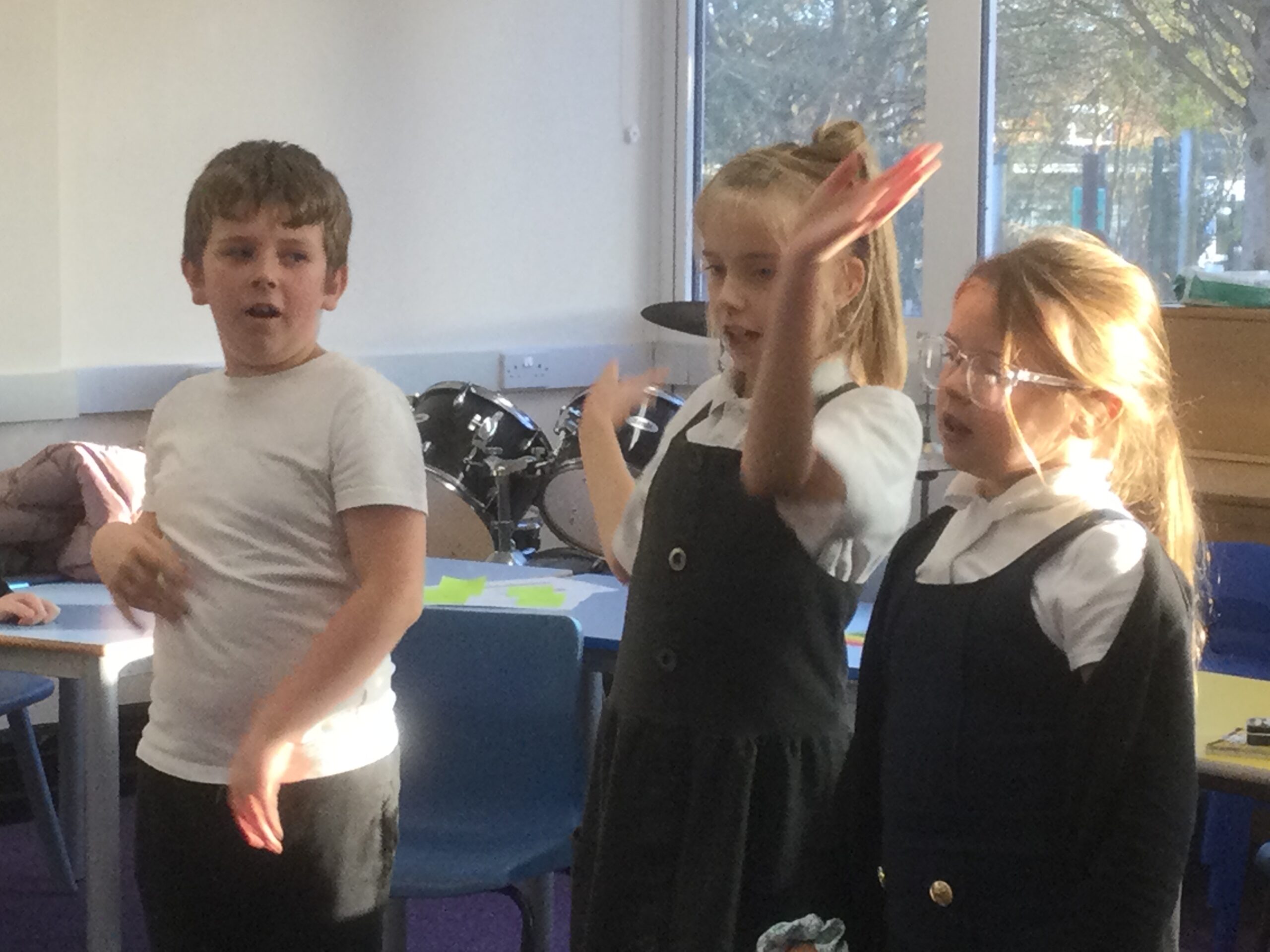 A row of two school girls and one boy, standing, the girl has her arms up in the air. Behind them is a drum kit