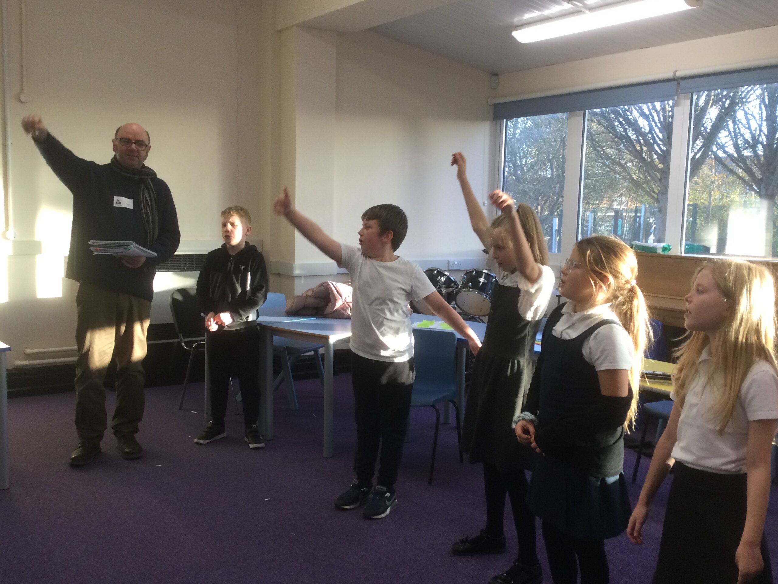 A row of school children standing in line in front of some tables, with a man, wearing a scarf, stands at one end - they all have their arms in the air