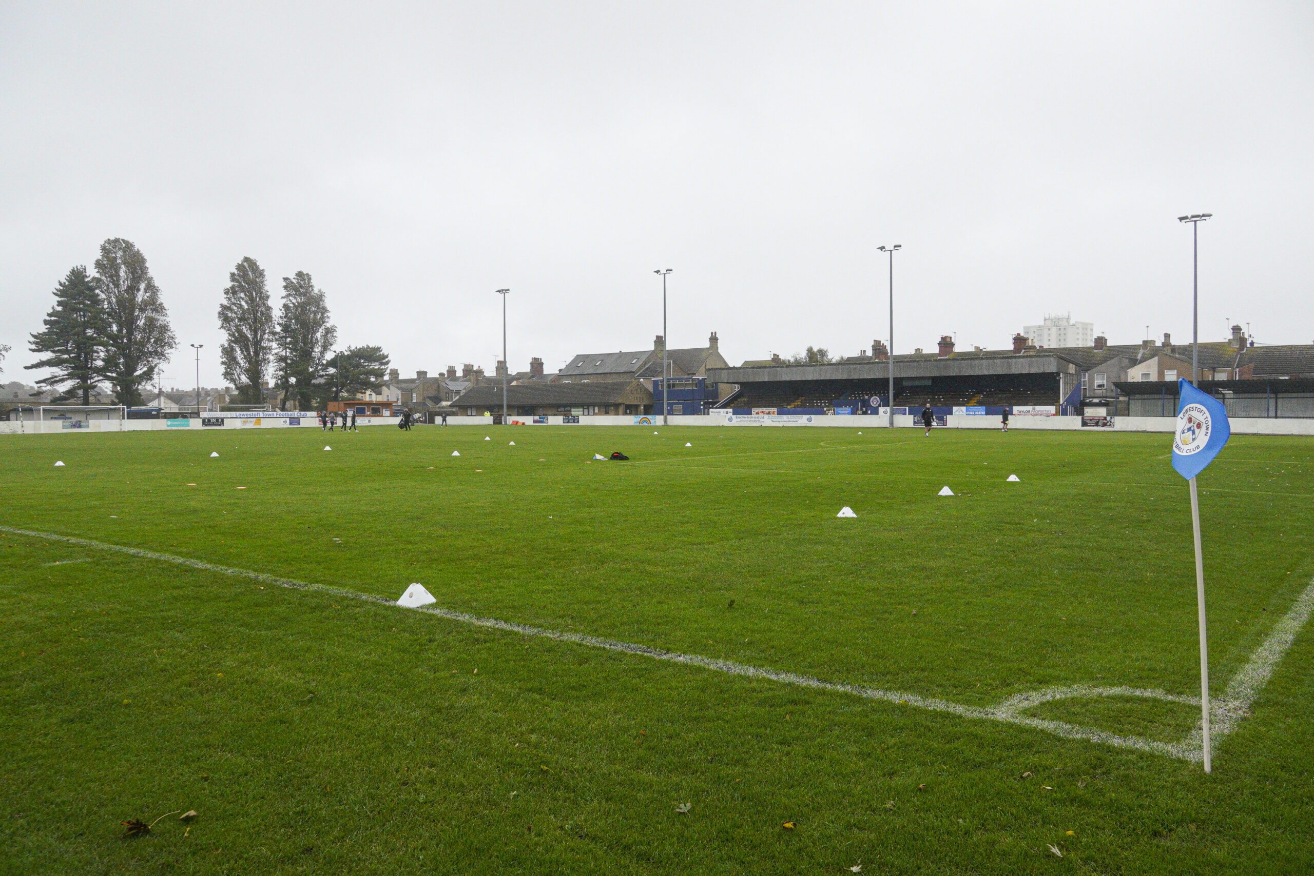 An empty football pitch with stands in the back ground. There are tall trees to the left of the pitch and white cones laid out as if in readiness for a training session. In the foreground is a white corner post with a light blue flag