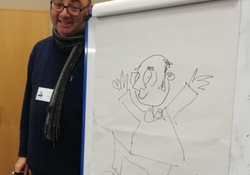A man wearing glasses stands beside a flip chart on which he has drawn a cartoon of himself