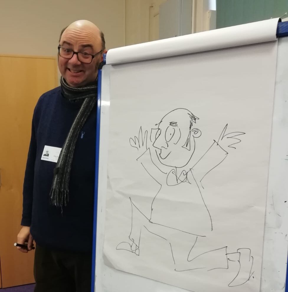 A man wearing glasses stands beside a flip chart on which he has drawn a cartoon of himself