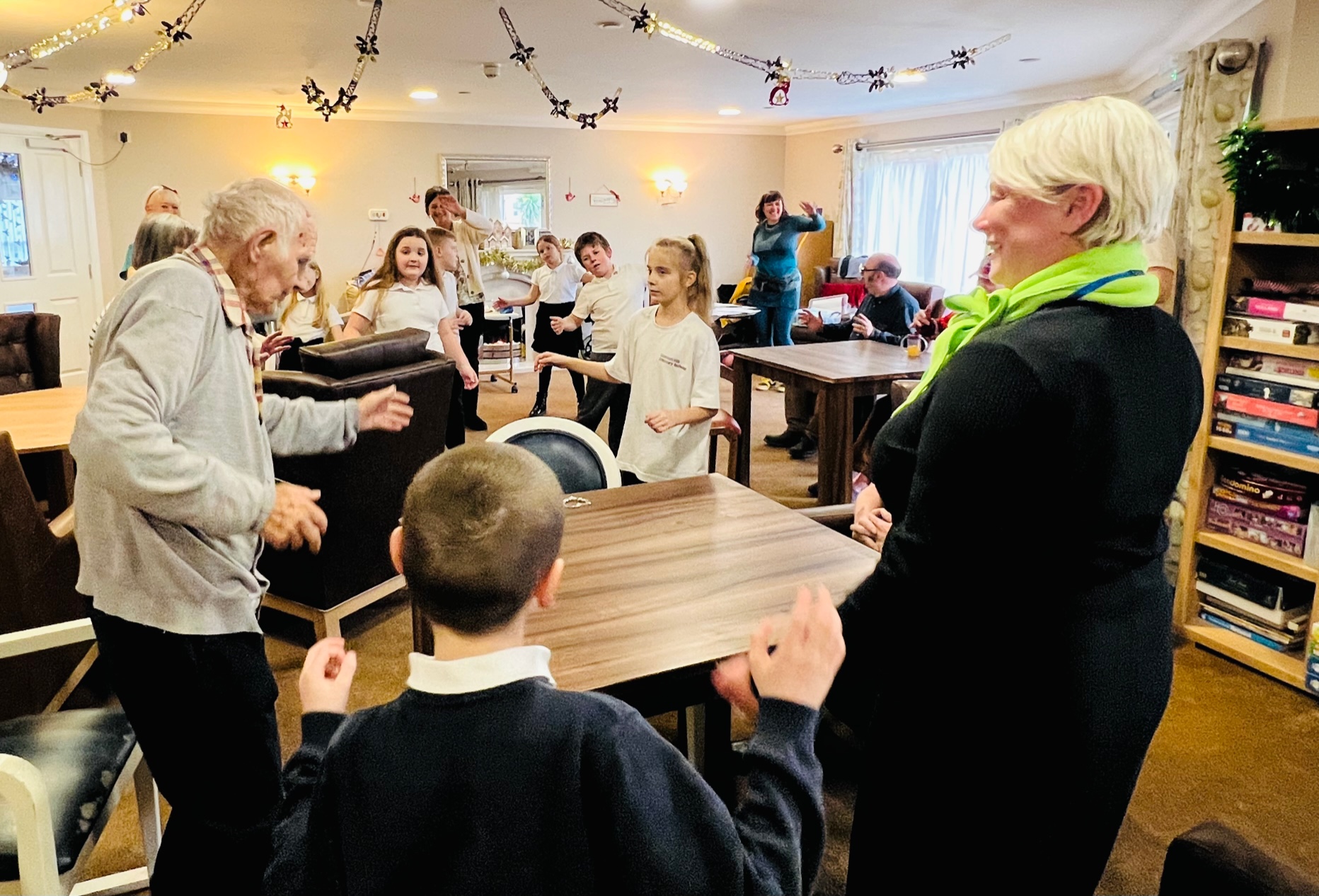 A gentleman wearing a grey cardigan and dark trousers stands with arms outstretched, leading the group in some exercises. Around the room are schoolchildren in black and white uniform and other adults with arms in the air. Foreground right stands a woman dressed in black and a lime green scarf