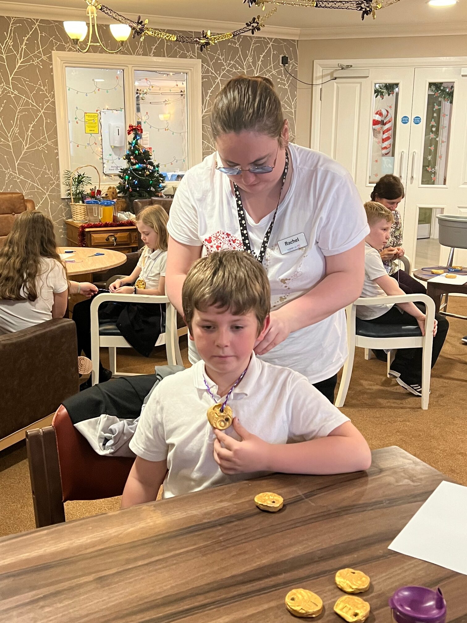 A woman stands behind a seated school boy, tying a ribbon around his neck whilst he supports a medal in his left hand. On the table beside him are 4 more gold medals