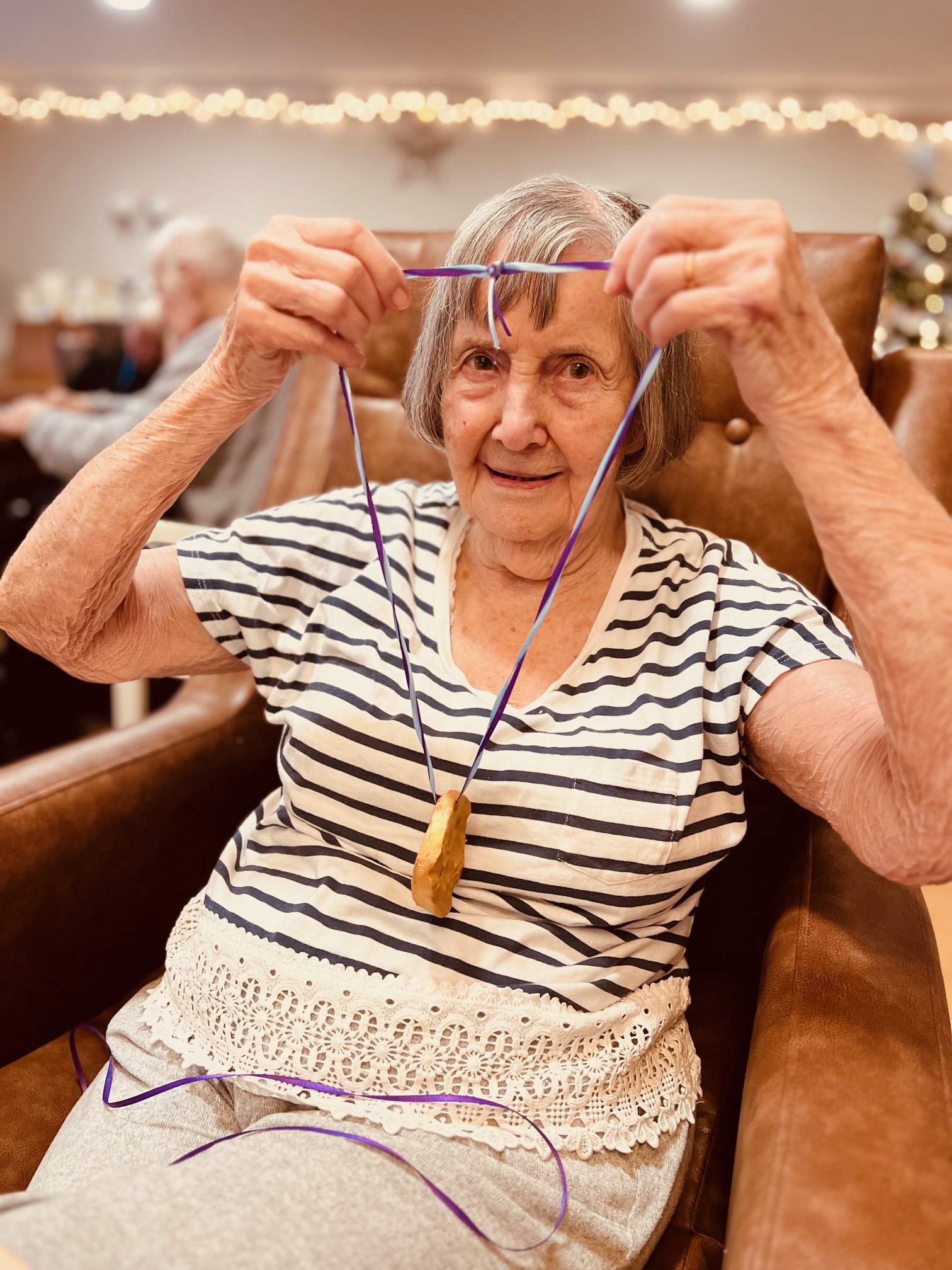 A woman wearing a striped top and seated in a brown armchair holds up her medal on a blue ribbon