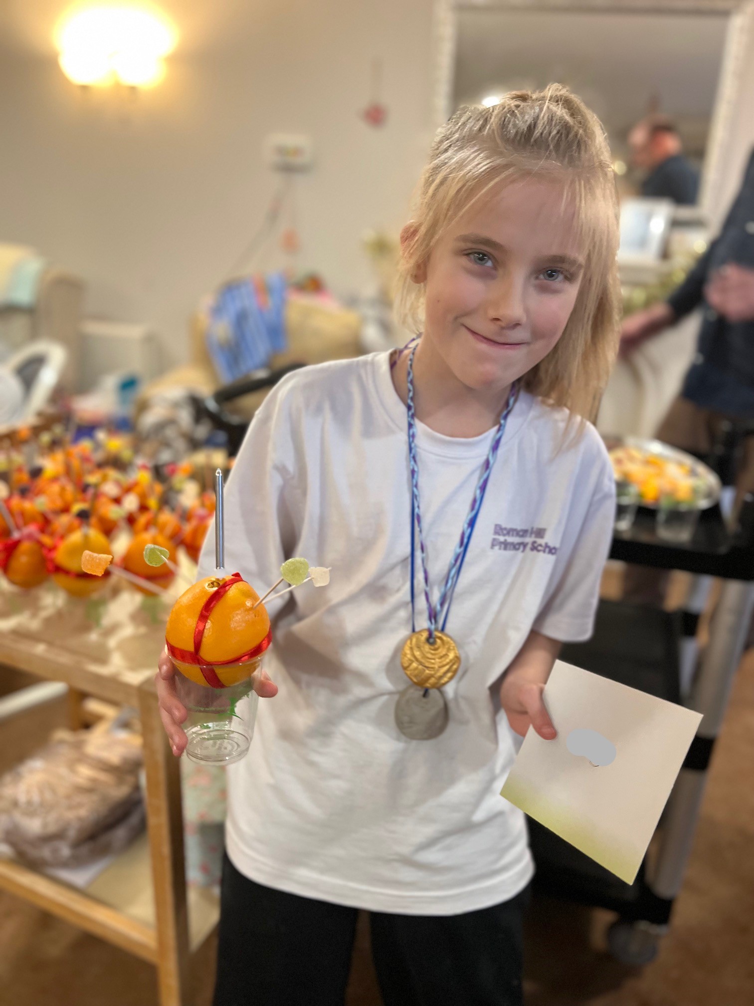 A school girl with blond hair tied back from her face, wearing a gold medallion and holding an envelope and an orange