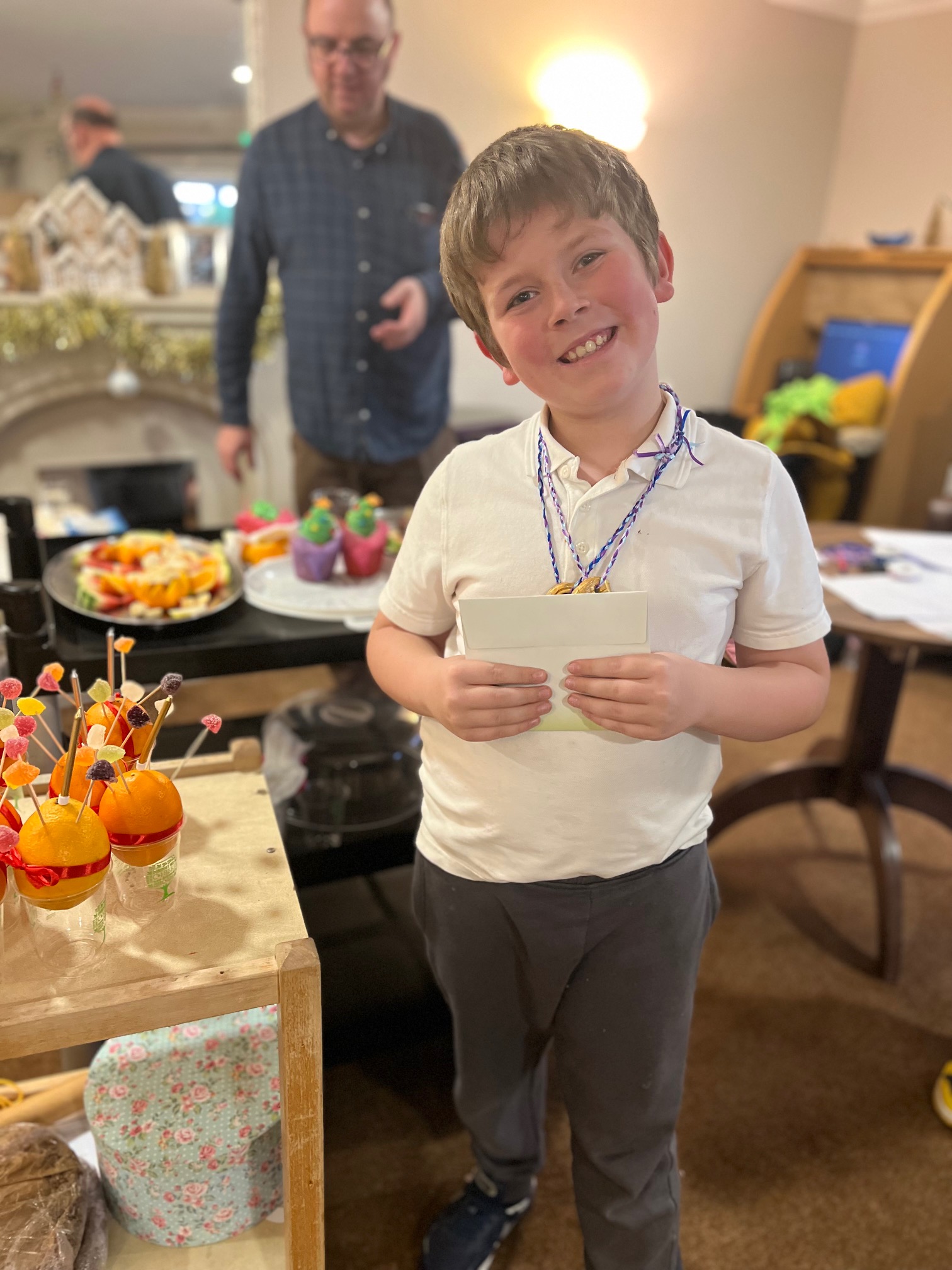 A boy faces camera, smiling, holding an envelope. On a trolley beside him are oranges, each balanced in a plastic cup, studded with sweets and a red ribbon tied around them