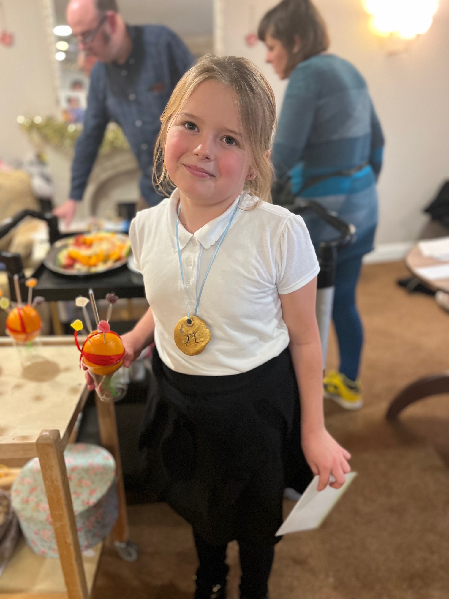 A child faces camera, wearing a gold medal around her neck on a blue ribbon and holding a card and an orange