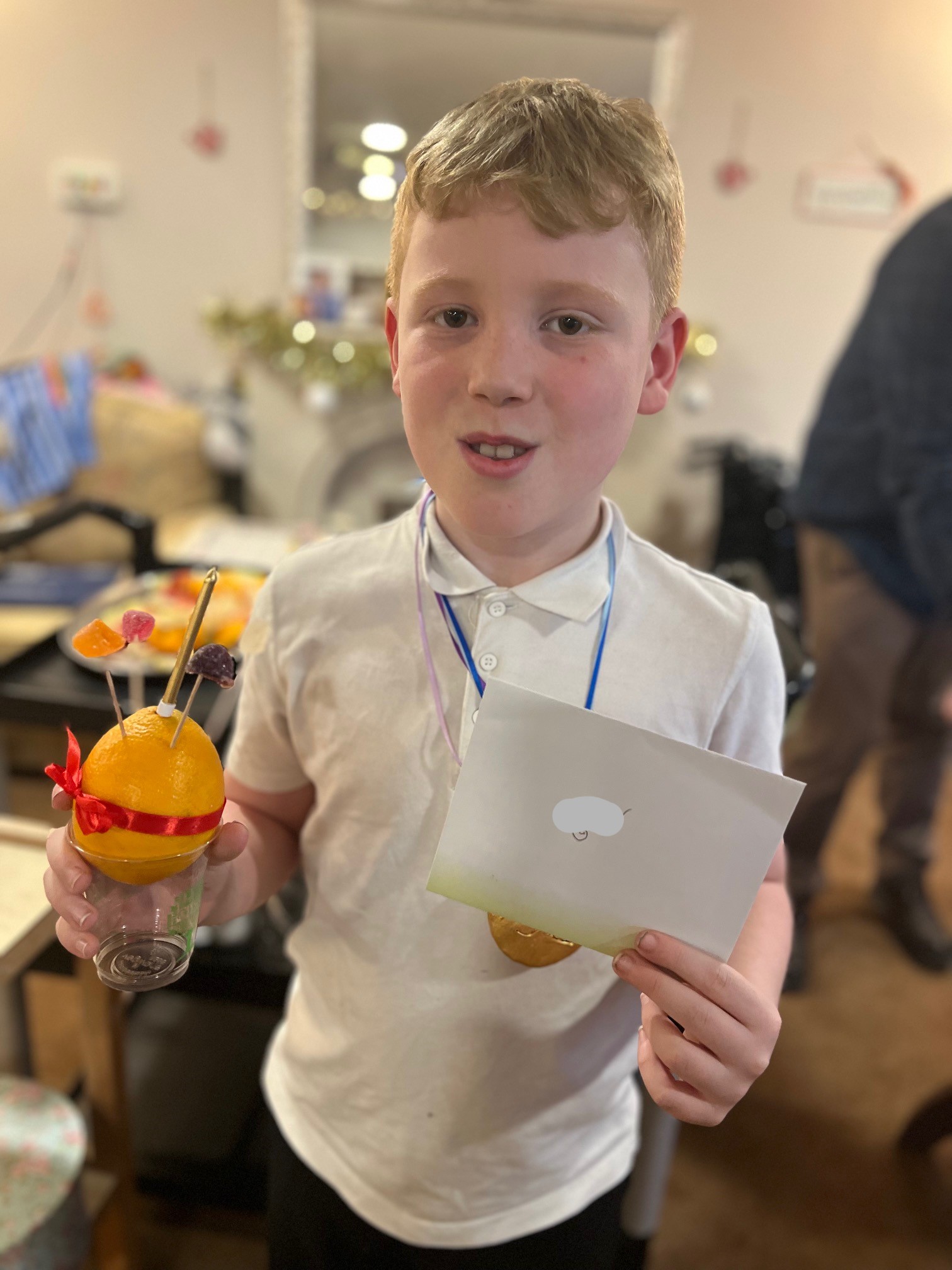 A boy faces camera holding an orange in a plastic beaker in one hand and an envelope in another
