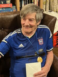 A gentleman in an Ipswich Town supporters top sits in a brown armchair. He has a medallion on his chest and is holding an envelope