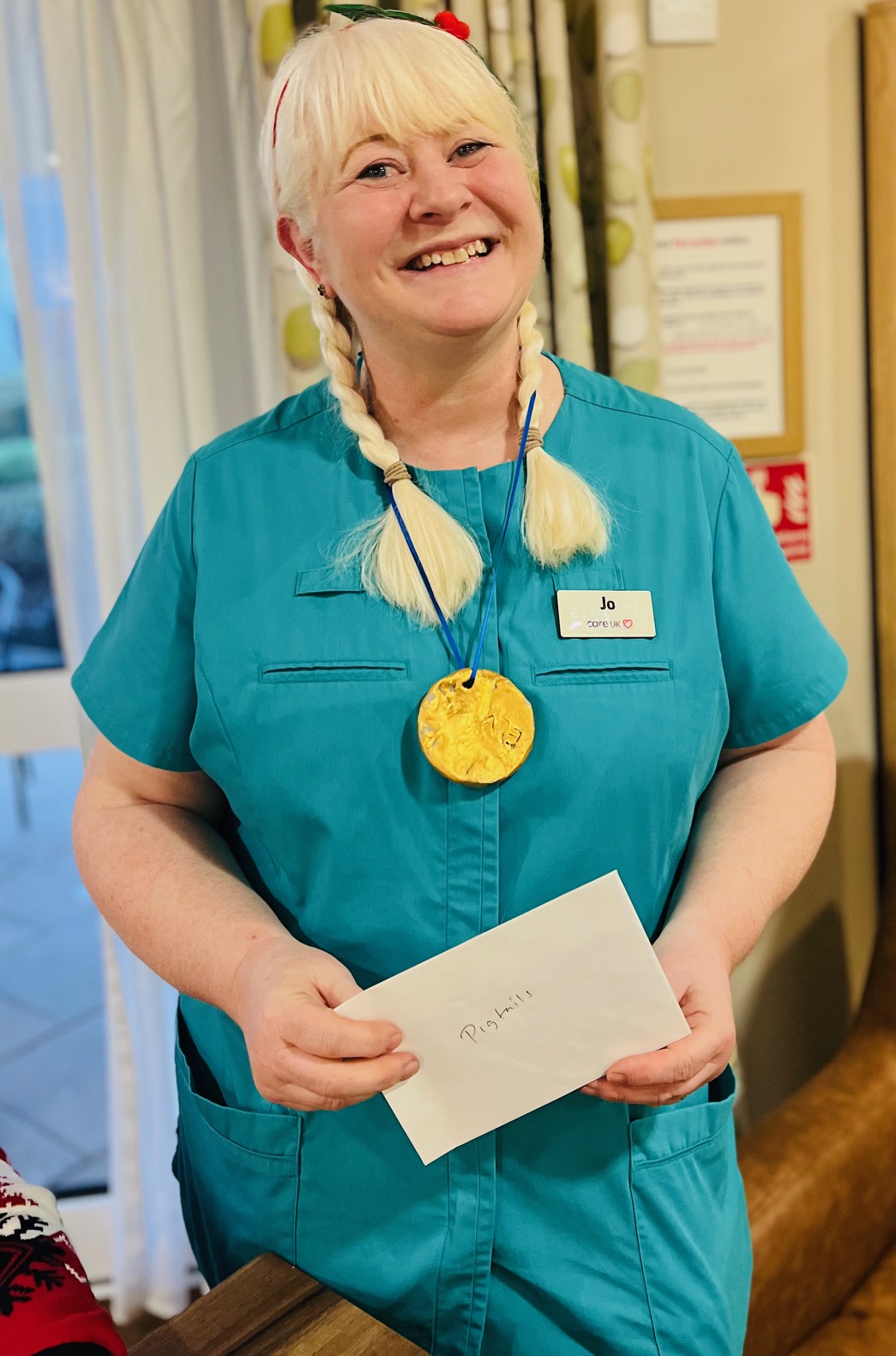 A woman in a turquoise top grins towards the camera. Her hair is braided in two pigtails that hang either side of her face and she is wearing a large gold medallion