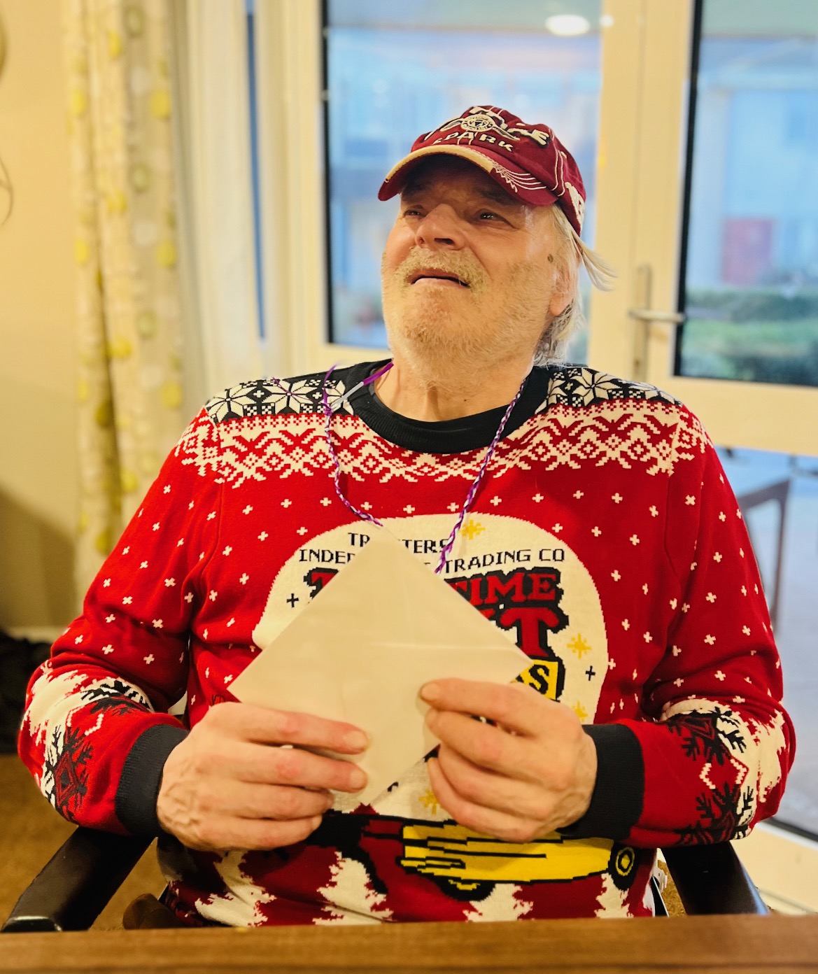 A gentleman wearing a red Christmas jumper and baseball cap, with a Christmas card in his hands