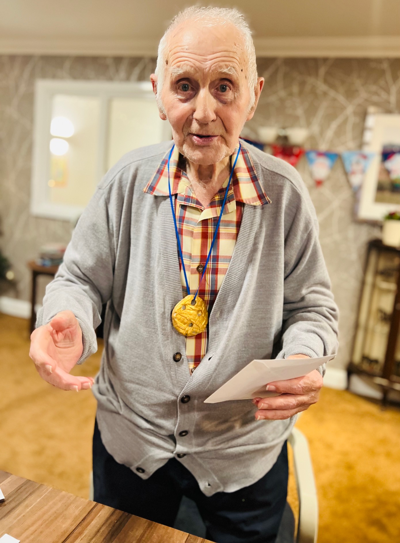 A gentleman wearing a check shirt and grey cardigan, with a gold medallion around his neck, looks to camera with his right hand outstretched