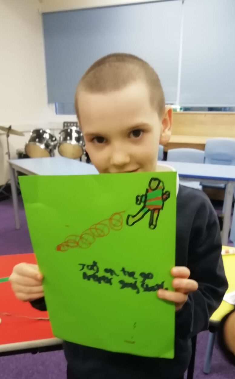 A schoolboy with a crew cut holds up a picture he's drawn of a footballer in a red and green striped jersey