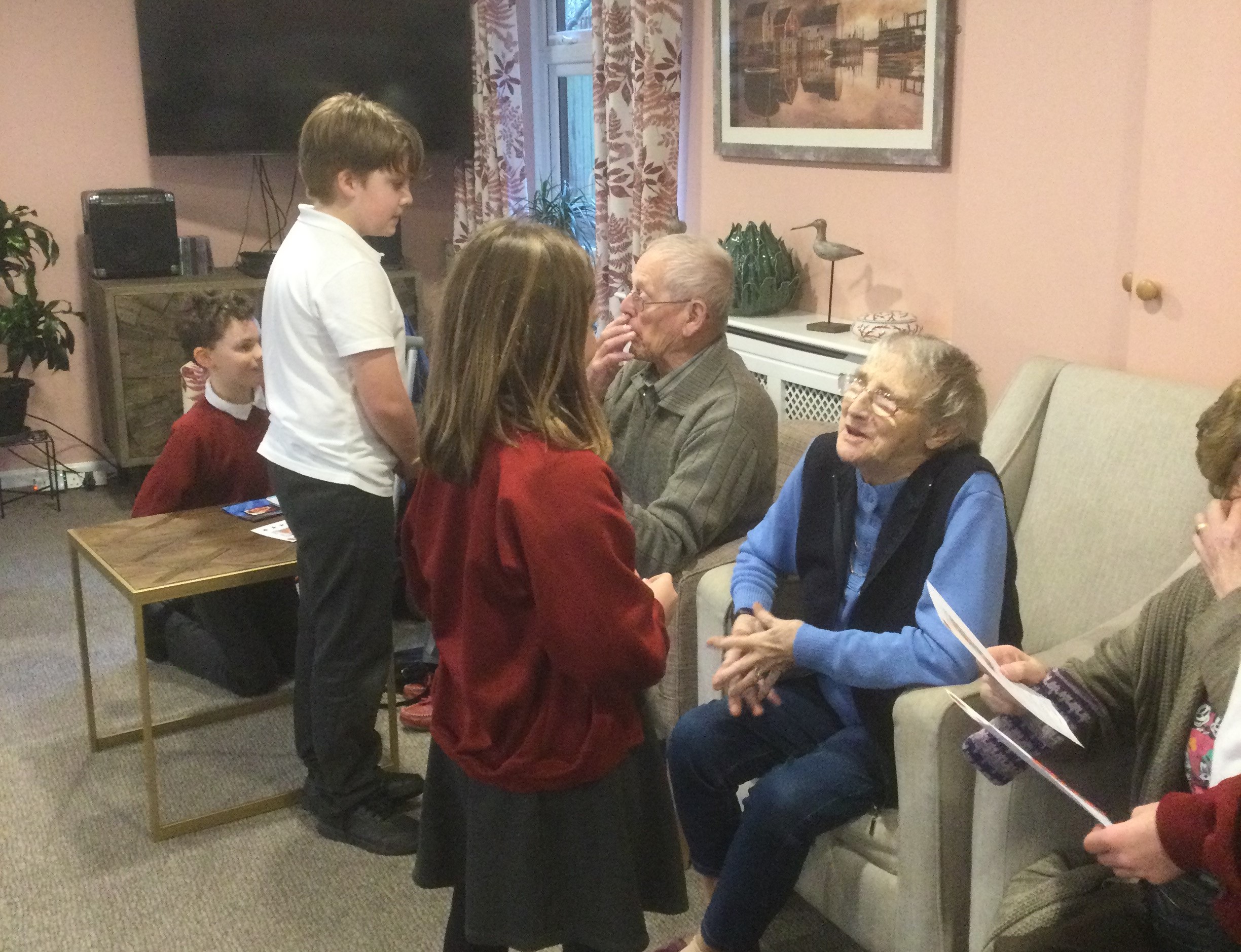 A row of school children standing in front of 3 adults, seated. One female adult smiles smiling at a child and all are clearly ddep in conversation