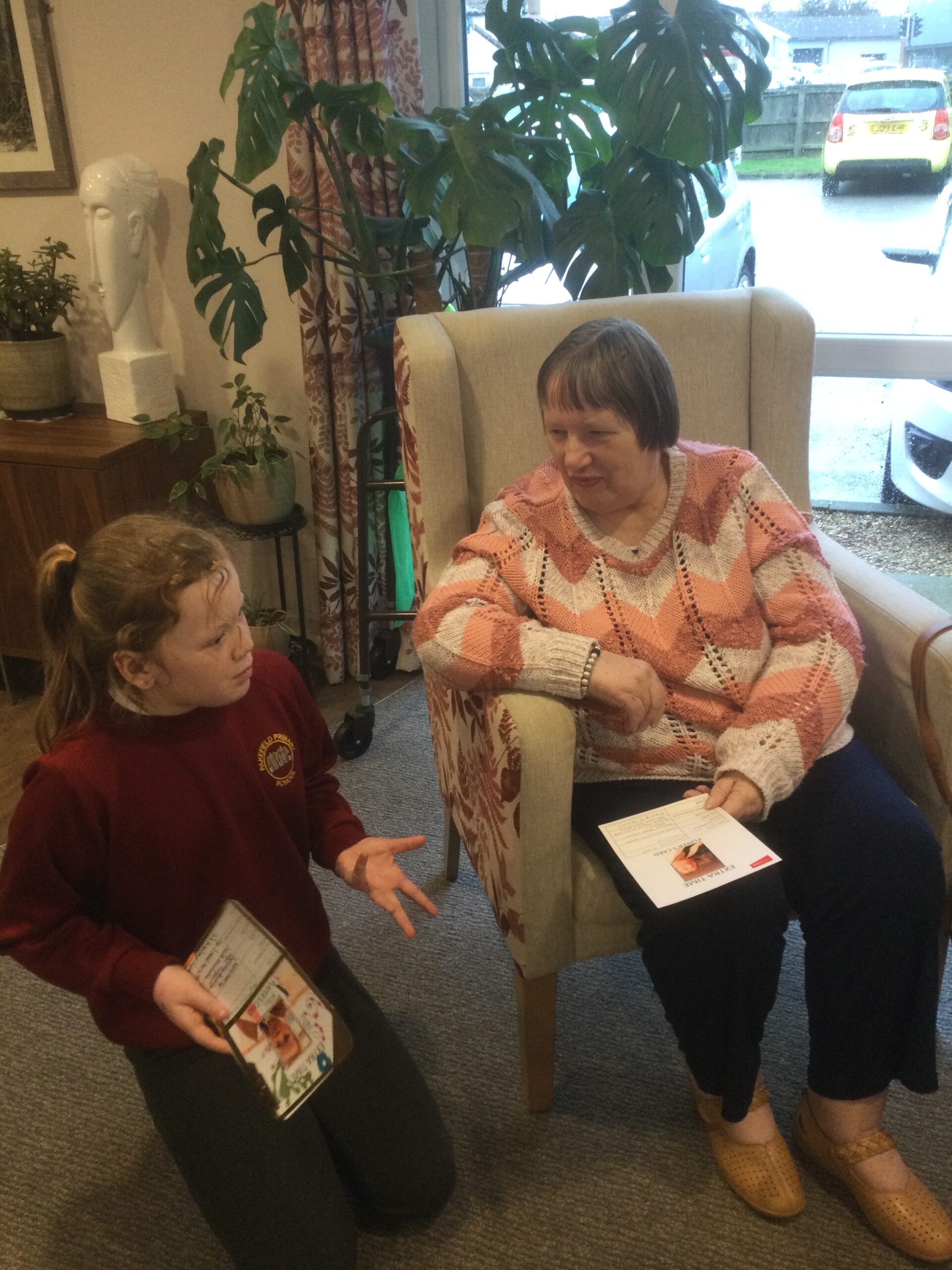 A school girl is kneeling beside a woman seated in an armchair. They are engaged in animated conversation