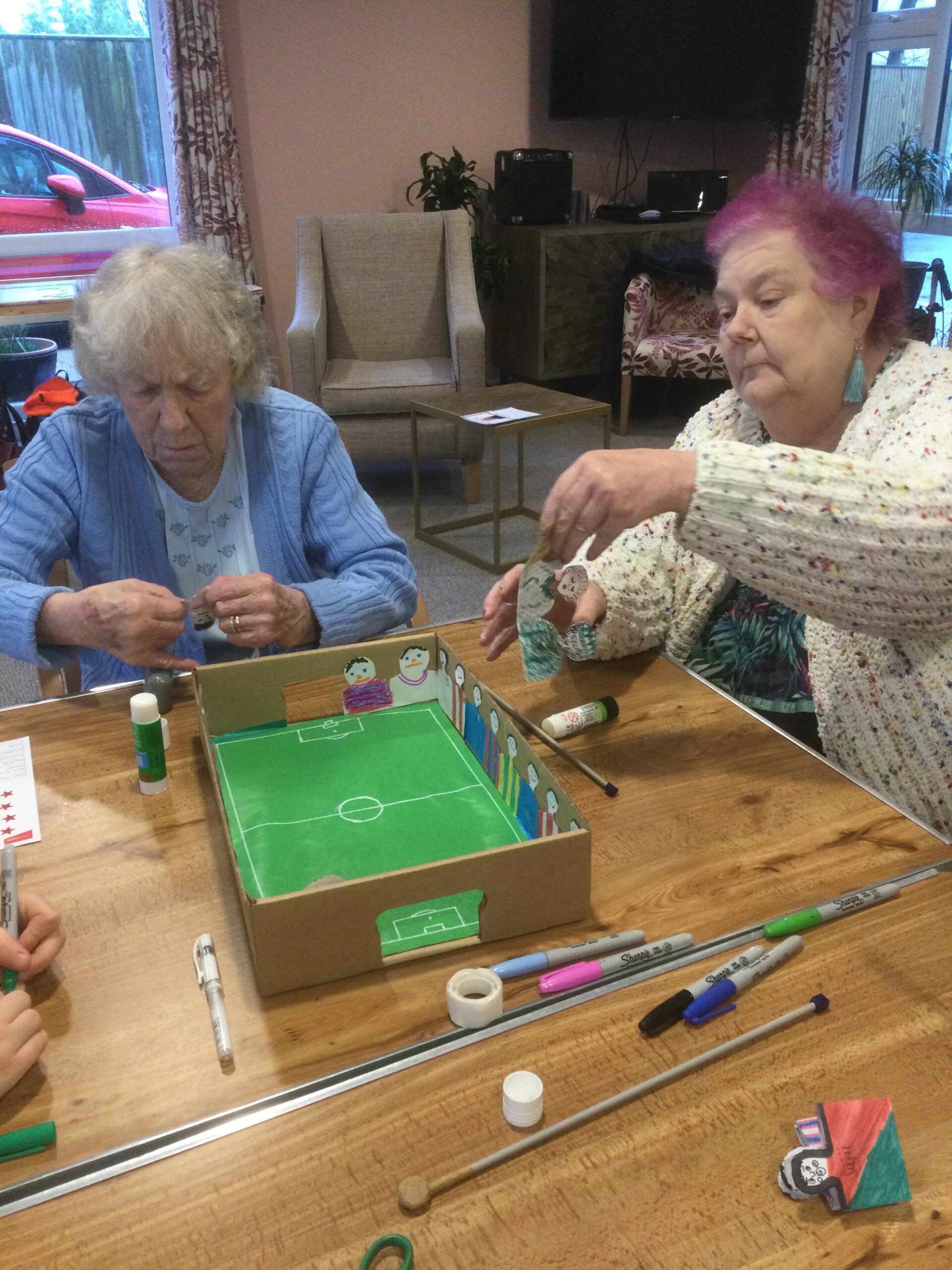 Two women seated at a table with a cardboard football game in front of them, pasting paper figures along the sides of the box