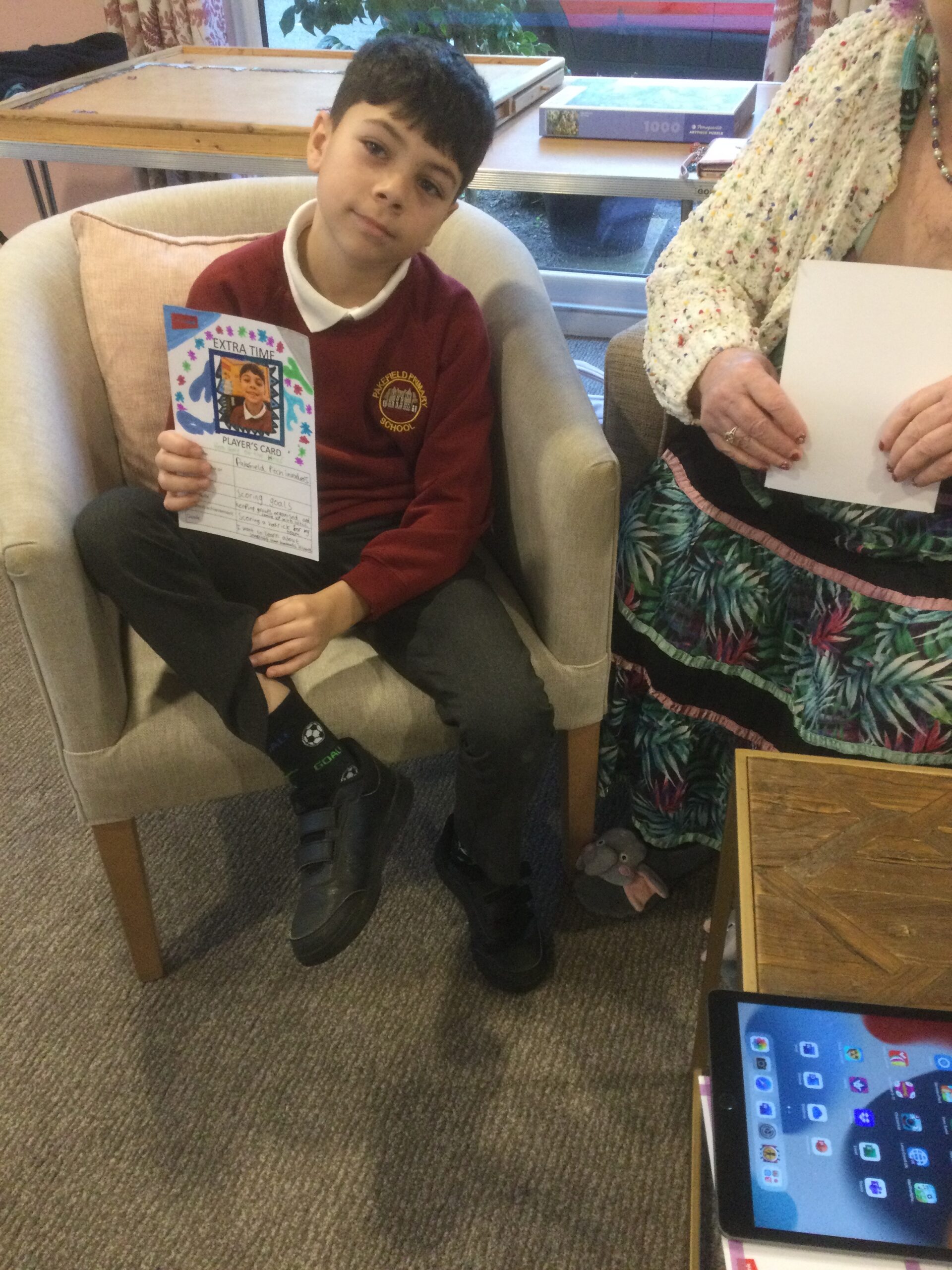 A school boy is seated in an armchair, facing the camera and pulling up a trouser leg to reveal a black sock with a football on it