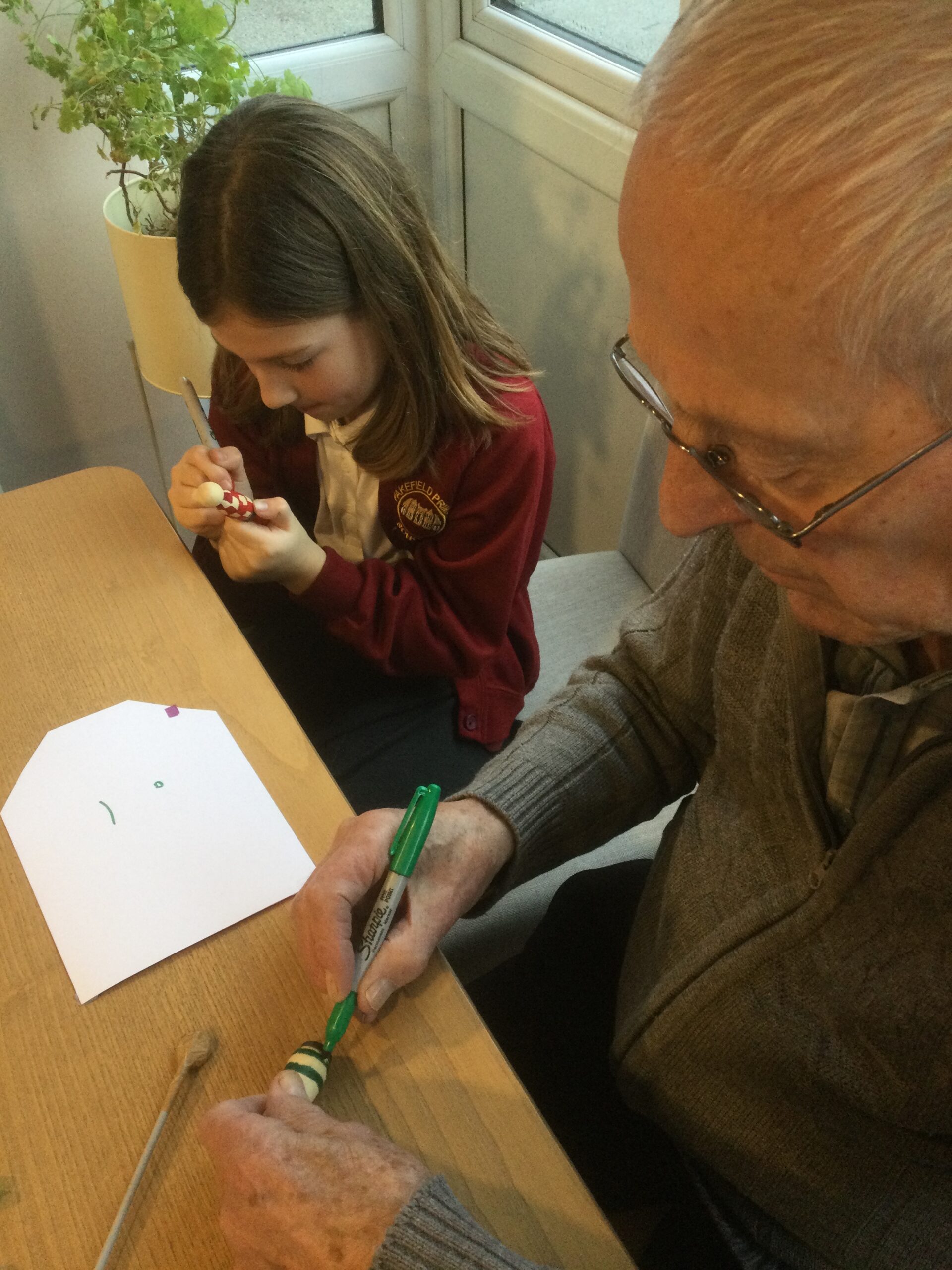 A school girl is seated beside an adult and both are fully focused on colouring a small wooden figure