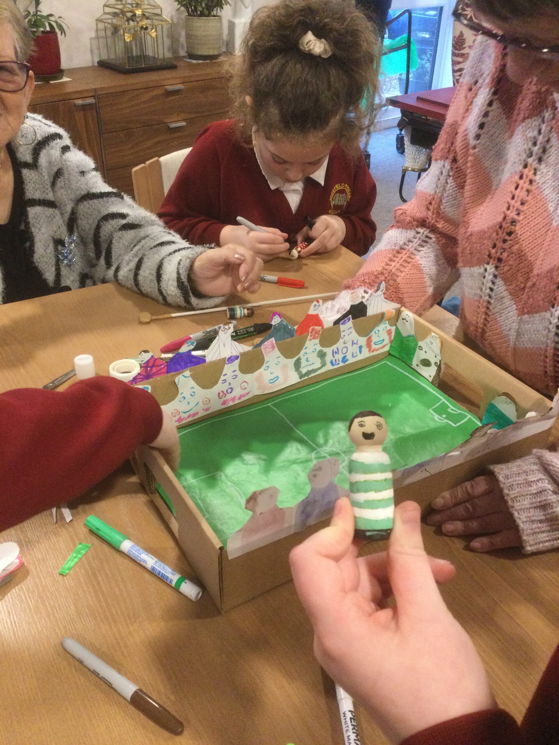 A group of adults and children seated around a table on which there is a cardboard football game. A hand holds up a small wooden figure decorated in green and white hoops