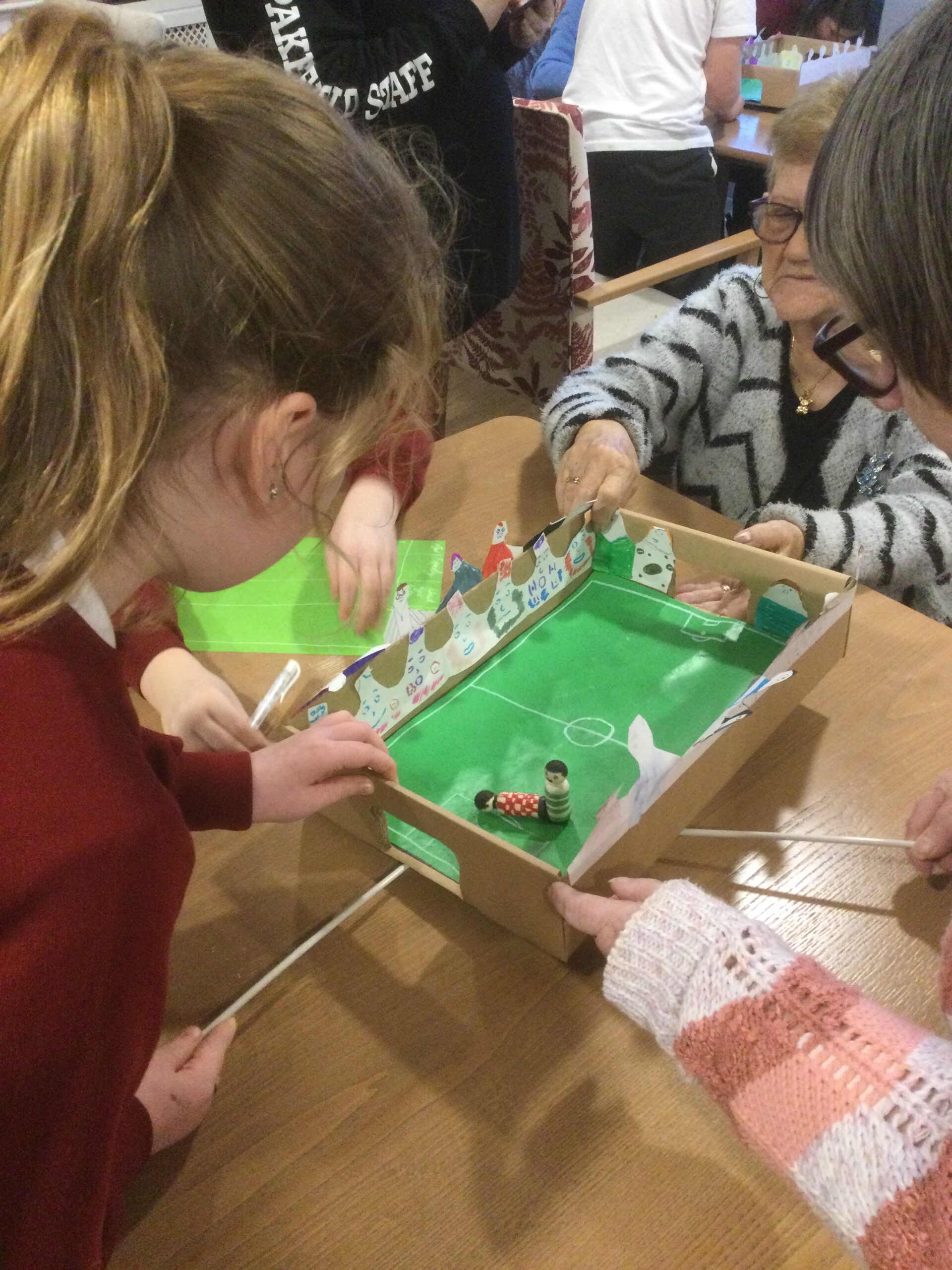 A cardboard football game is on the table. An adult and a child each has a stick running under the box, used to propel the magnetic football figure across the board. Another adult steadies the game