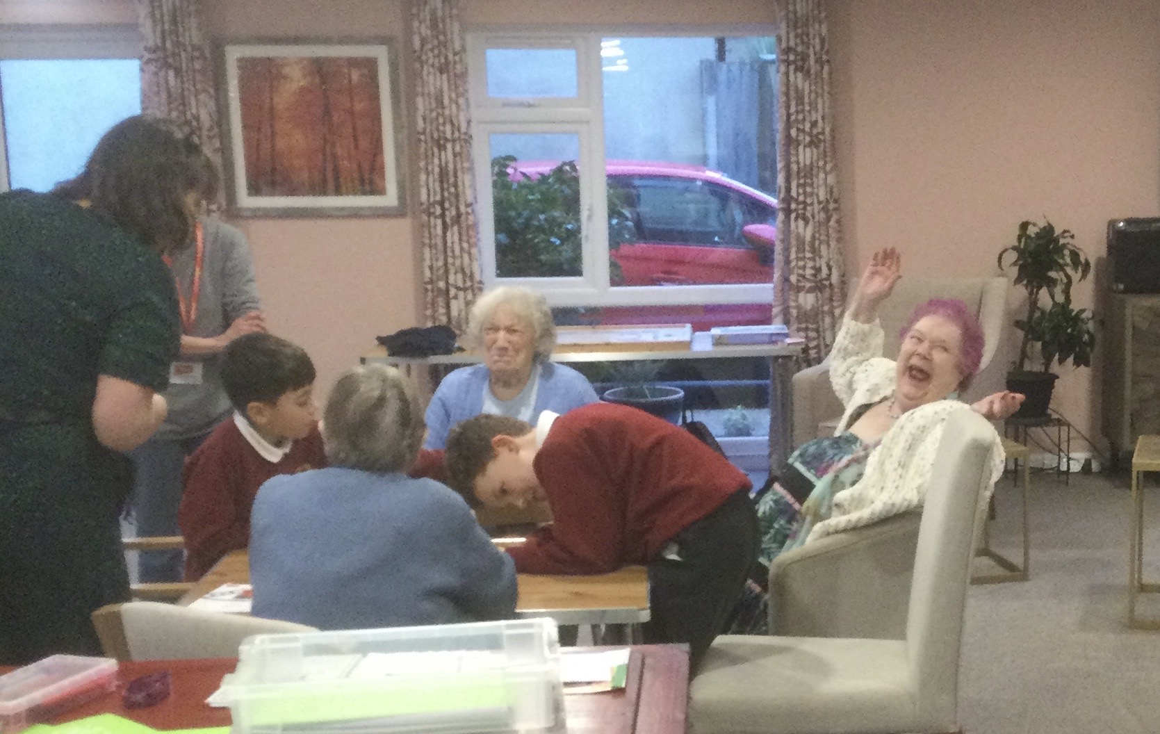 A woman leans back in her chair, hands in the air, mouth wide open in a huge grin, clearly pleased about something. Another woman beside her grins as others look on at what they're playing on the table