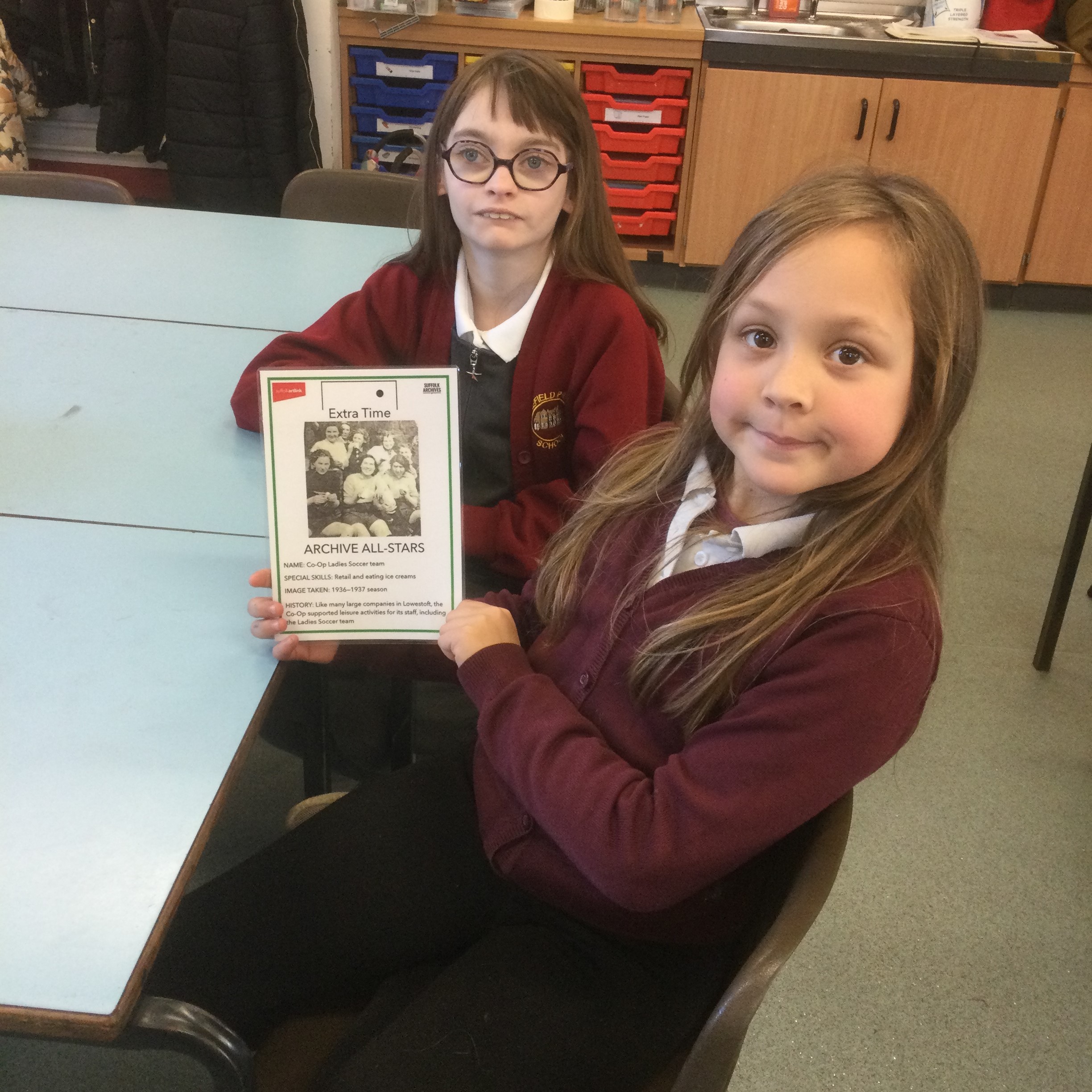 Two school girls seated at a blue table, holding up a plasticised card with a picture of a ladies football team on it